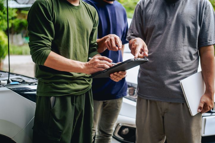 Asian clients engage with a car insurance agent beside a wrecked vehicle. They examine the policy details, assess the damage, and finalize paperwork related to the insurance claim and repairs.